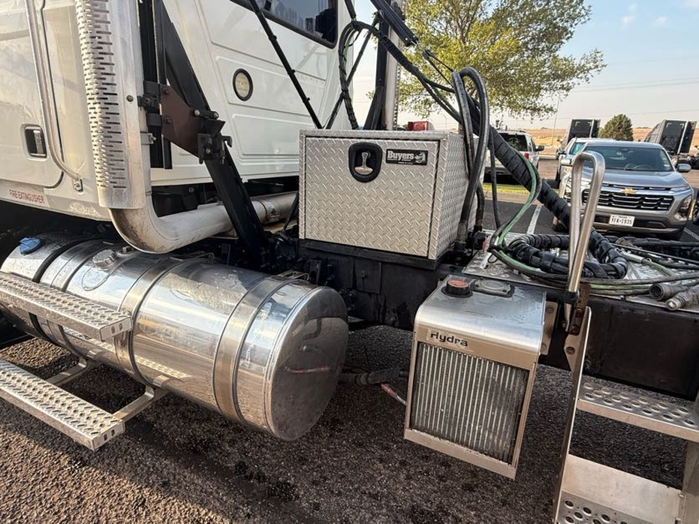 Mack Pinnacle truck cab area showing a chrome cylindrical fuel tank, aluminum toolbox, Hydramatic or Hydrive system with fuel lines and steps; metallic panels and hoses visible.