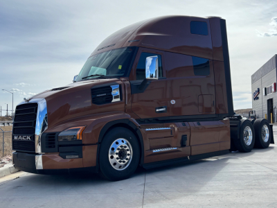 Mack PR64T76H truck tractor in brown shown in side-front view with sleeper cab and chrome mirrors.