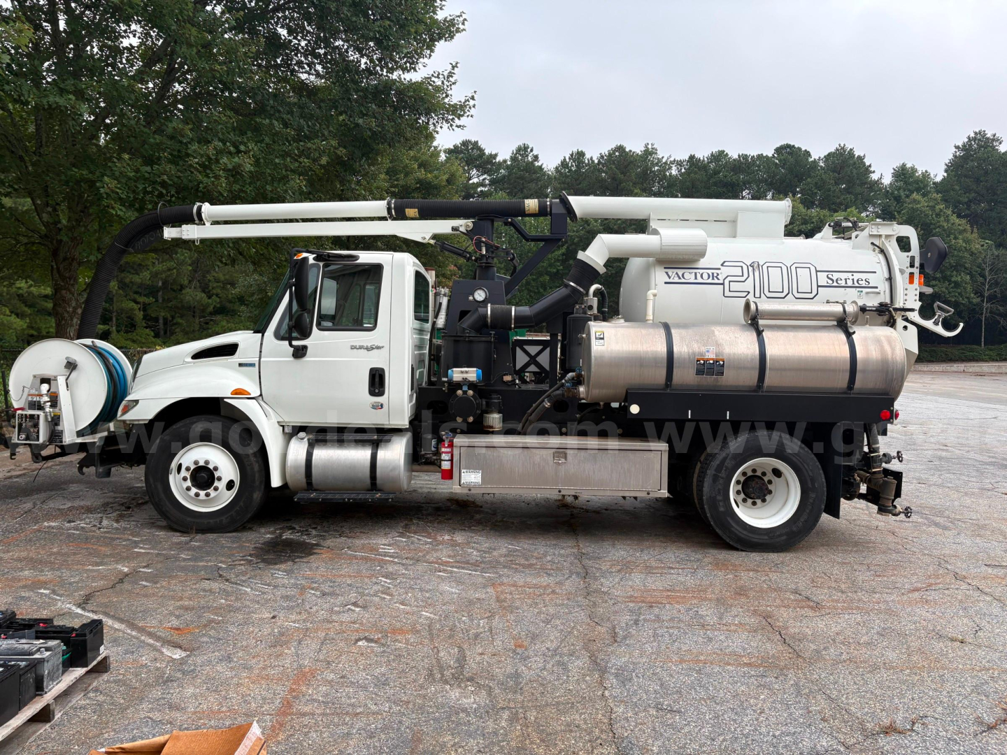 Side view of a white International 4300 vacuum truck with a large stainless tank, pump, and hose reel. Visible text includes '2100 Series' on tank and 'Victor' branding, hoses, and control panels.