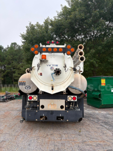 Back view of a vacuum truck tank with two large rear hoses on the right, multiple orange lights, rear bumper, and a circular tank hatch labeled VACTOR.