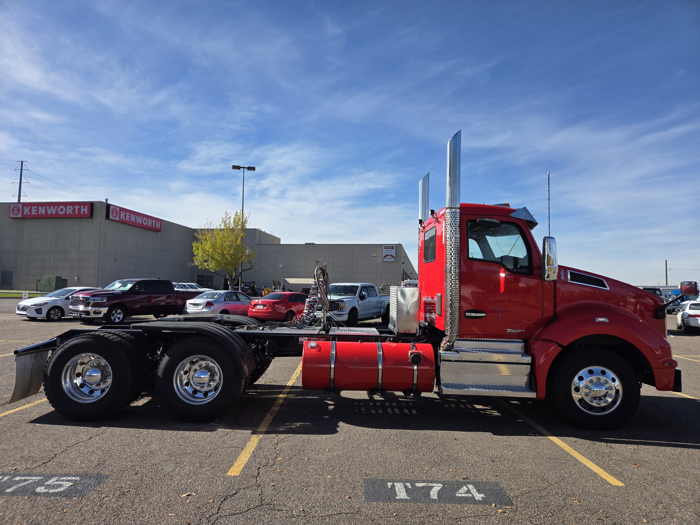 Kenworth T880 conventional daycab truck side view with red cab and chrome exhaust stacks; visible are the fuel tanks, wheels, and a semi-truck yard in background.