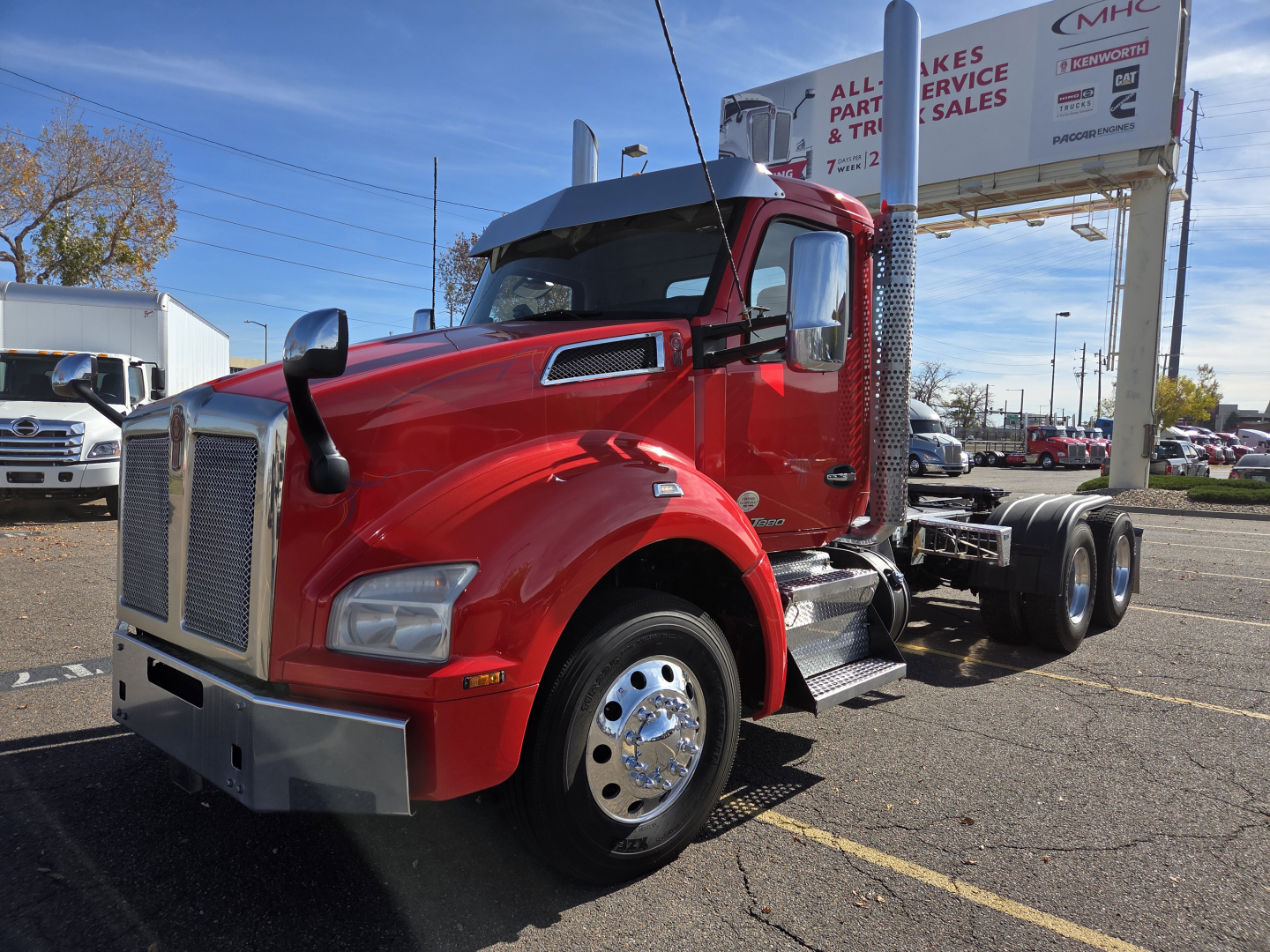 Kenworth T880 conventional daycab truck in red with chrome bumper and large grille; side mirrors; dual exhaust stacks; text on signs in background.