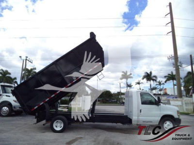 Truck bed/dump dump-box in raised position on a white Ford E-450 commercial truck, with front cab visible; stock yard background with palm trees.