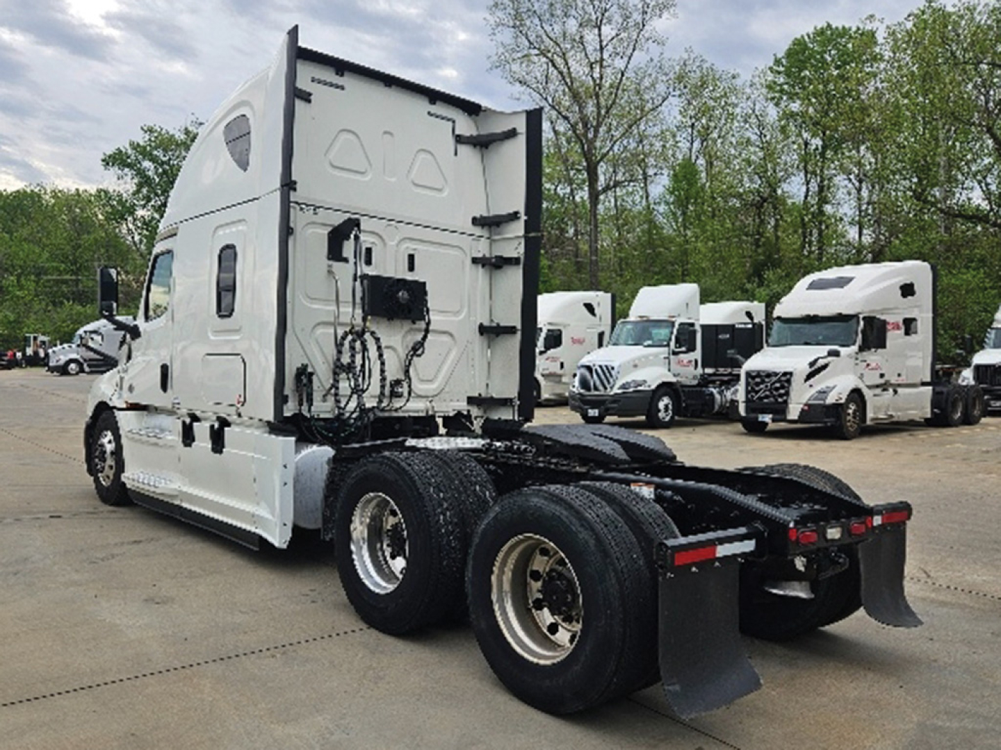 Truck chassis of a 2020 Freightliner Cascadia126 sleeper cab seen from rear three-quarter view; dual rear axles, fifth-wheel area visible, white paint, multiple trucks in background.