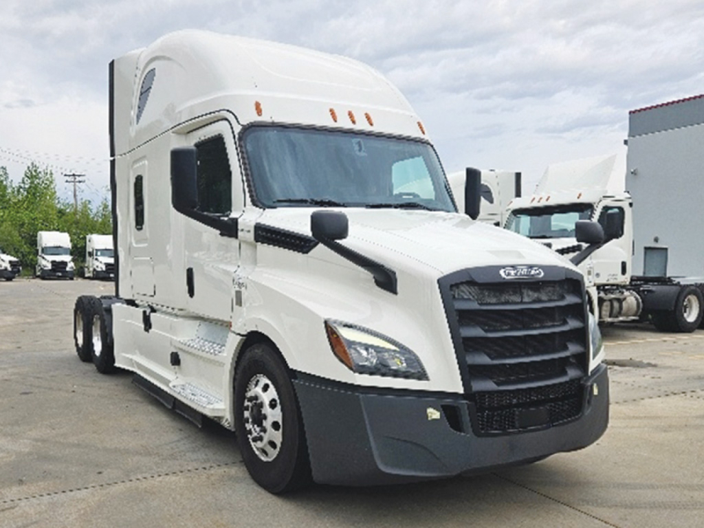Semi tractor truck in a lot showing Freightliner Cascadia interior details; front grille with black plastic, large windshield, side mirrors, and cab steps.