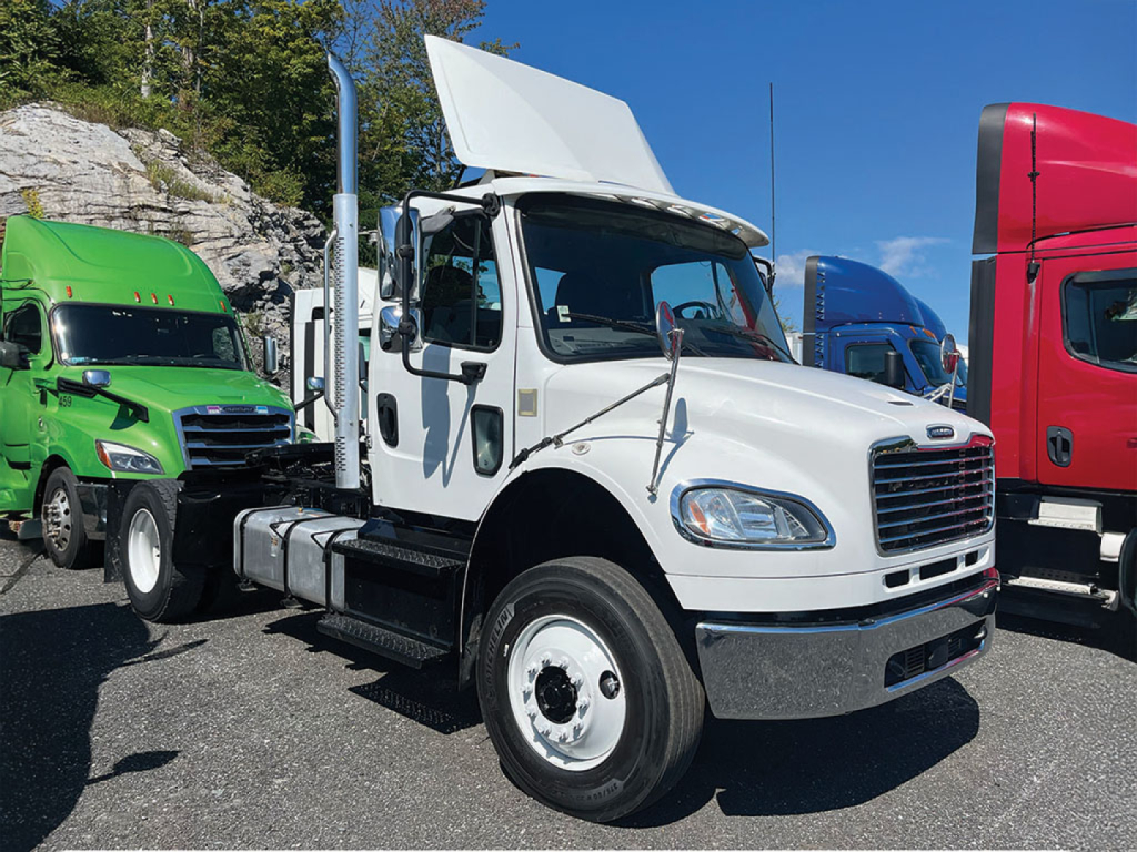 White Freightliner M2 106 daycab truck with hood open, parked among other colored trucks; side mirrors, exhaust stack, and front grille visible.