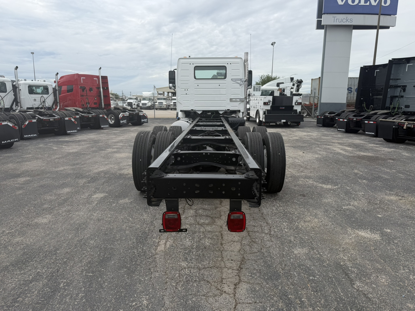 Rear view of a white Volvo cab-and-chassis truck frame showing dual rear axles, chassis rails, and taillights. An open truck yard with other trucks and a Volvo sign in the background.