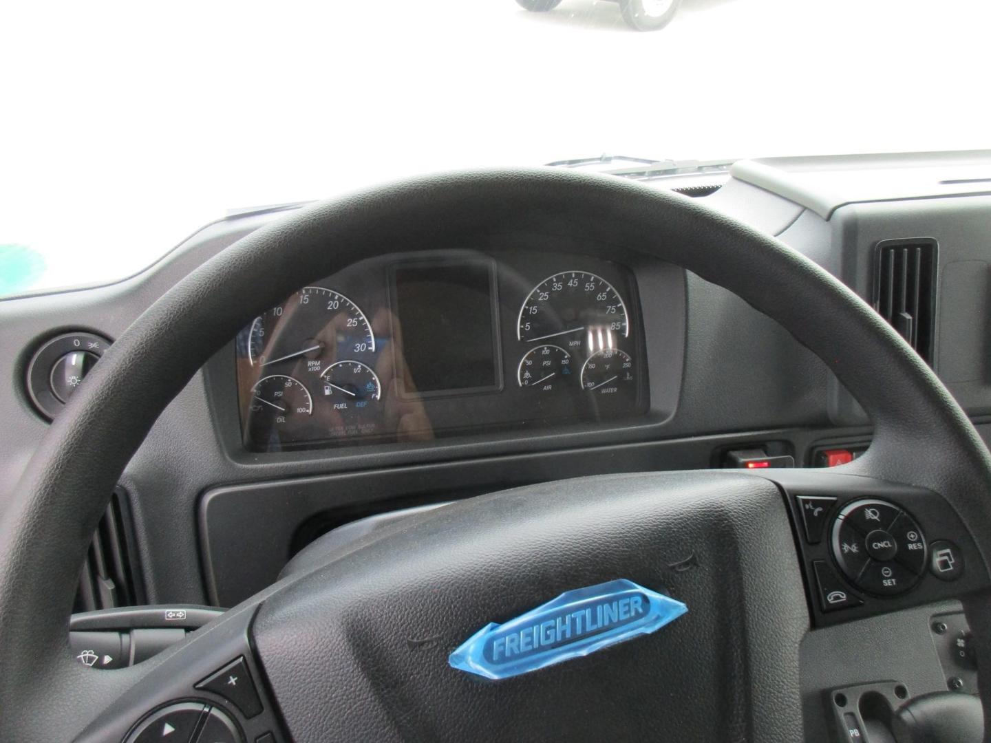 Dashboard and steering wheel of a Freightliner truck. Visible text on the emblem reads 'FREIGHTLINER' on the blue badge. Instrument cluster with gauges and digital display.