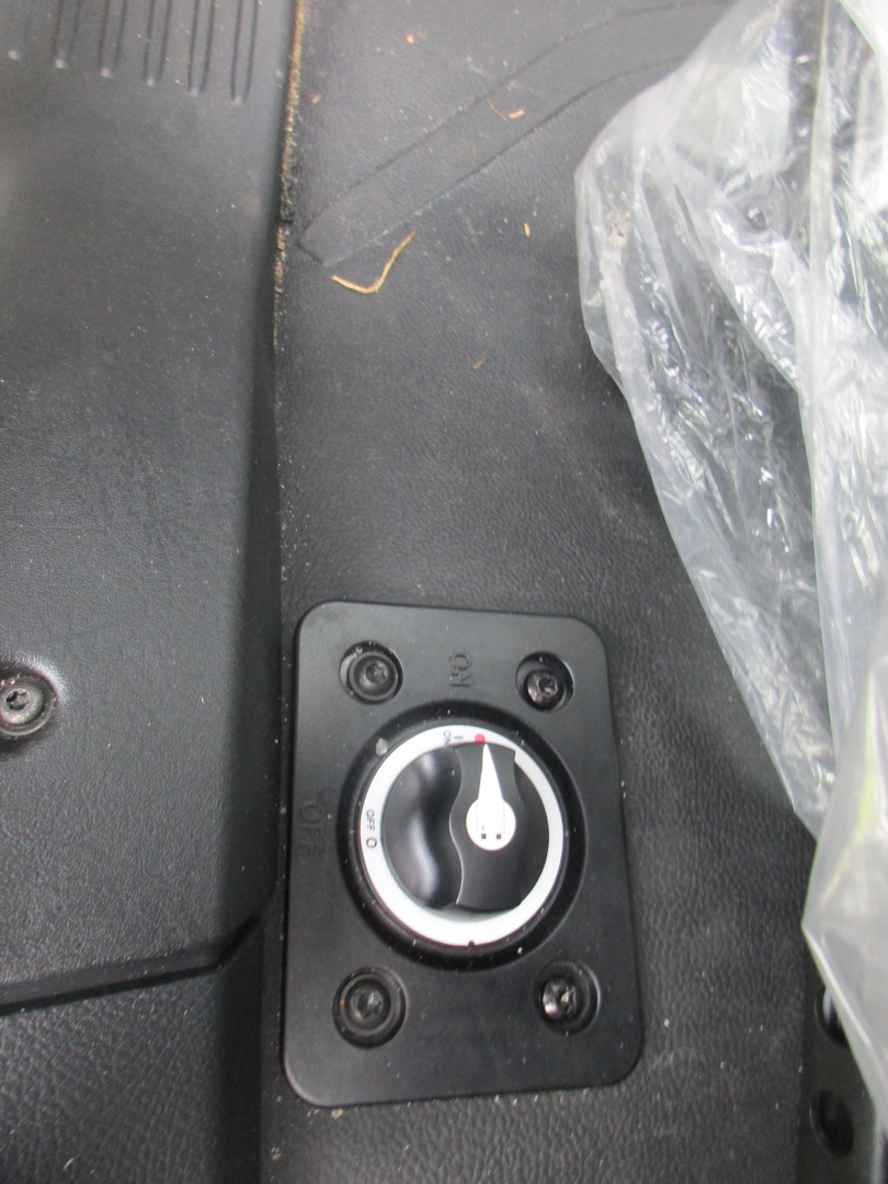 Vehicle switch panel with ignition-style dial inside a rectangular mounting plate, surrounded by screws, located on a black textured surface.