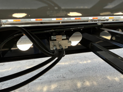 Trailer undercarriage with hydraulic lines connected to a metal valve block on a black steel frame. Visible hoses and mounting hardware beneath the side dump trailer.