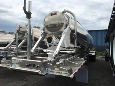 Heil Cementer trailer with polished stainless steel tank showing Heil brand label; multiple support frame arms and hoses; control panel with labels on the front; reflective surface; trailer chassis with red-white safety stripes.