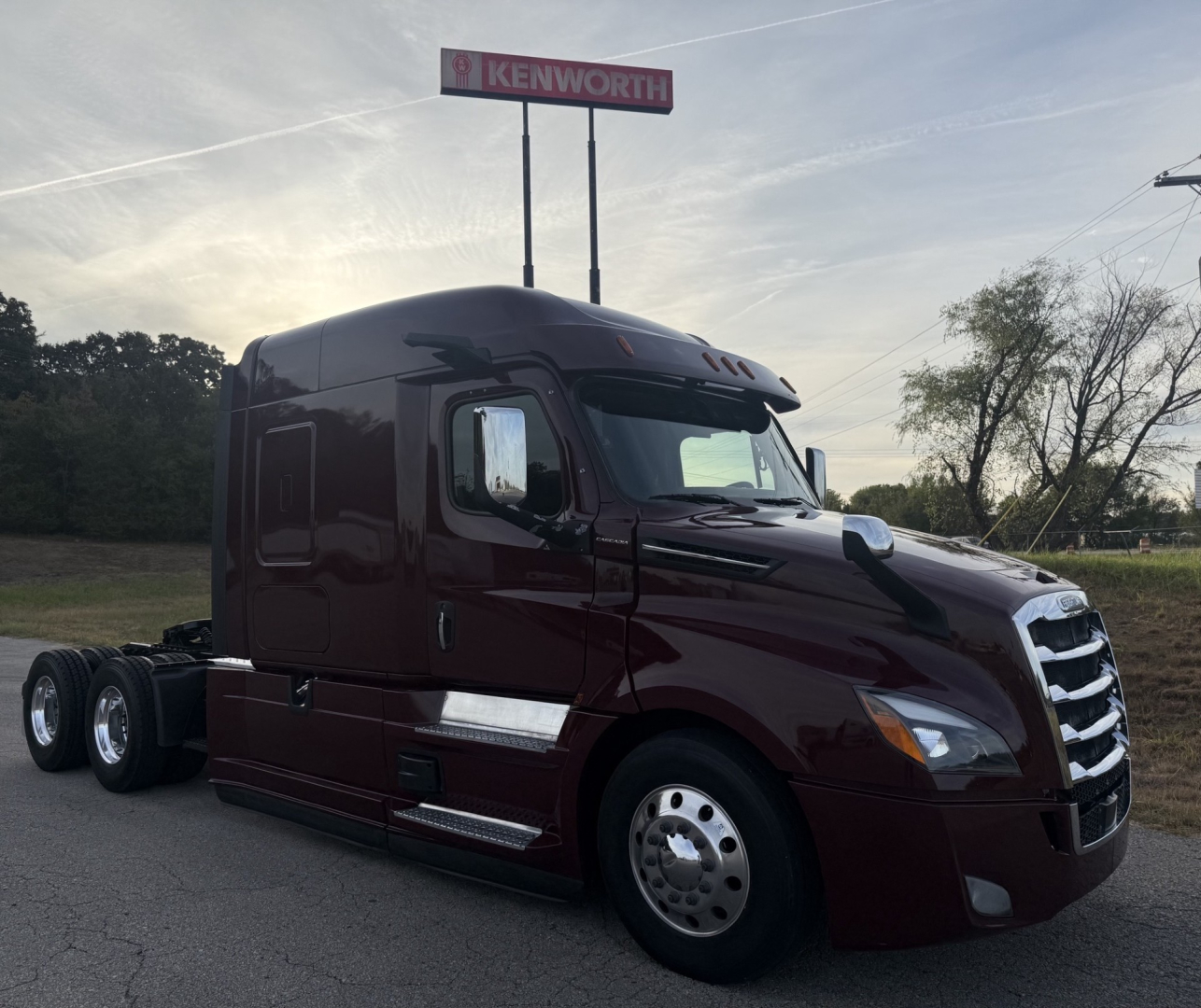 Truck cab with maroon Freightliner Cascadia 126 semi-truck showing side mirrors, chrome fuel tanks, dual rear axles, and Kenworth sign in distance.