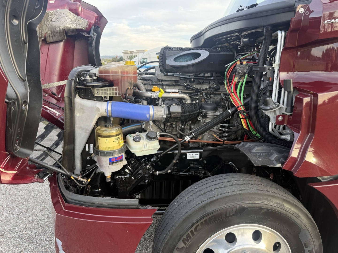 Engine bay of Freightliner Cascadia126 with exposed components including air intake hose, reservoirs, and wiring; blue hose visible.