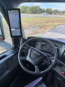 Cab interior of a 2021 Freightliner Cascadia 126 with steering wheel, dashboard digital gauge cluster, and side mirror selfie screen.