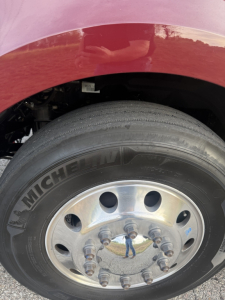 Truck wheel and tire close-up showing Michelin tire on front dual-axle rim with lug nuts and part of red fender.