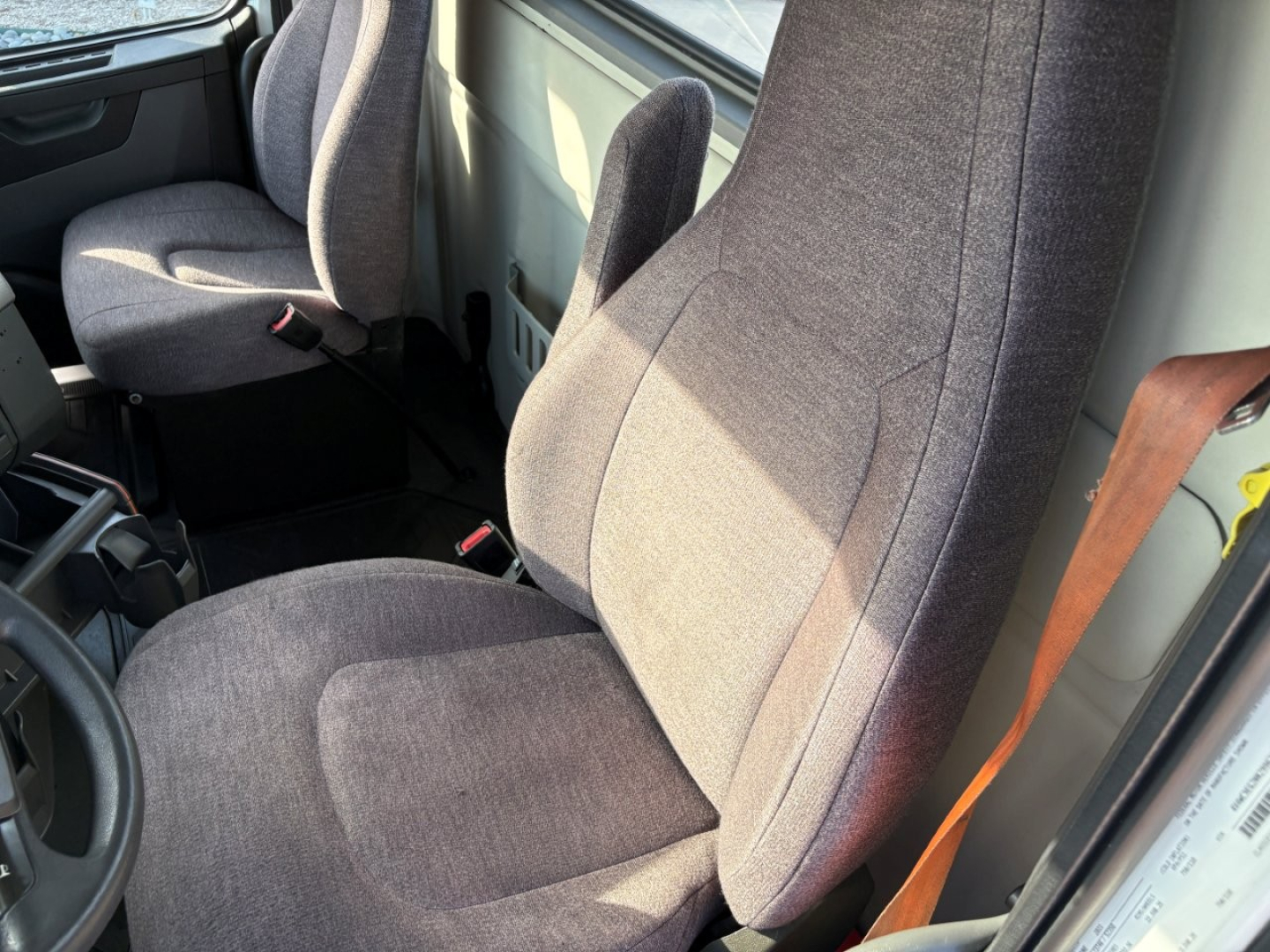 Front cabin interior of a 2022 Volvo VNR64T300 showing driver seat and passenger seat with gray fabric upholstery and seat belts.