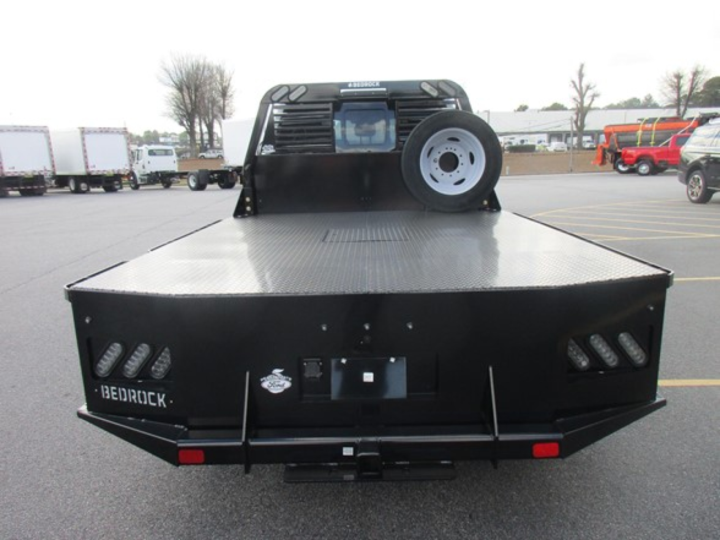 Flatbed truck in rear view showing bed surface, spare tire mounted on cab-back, and dual rear lights with diamond plate surface.