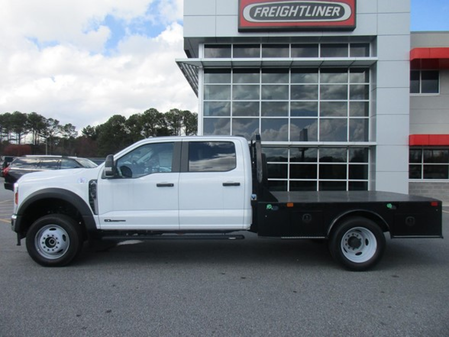 Ford F-450 flatbed truck side view in front of dealership, white cab, black flatbed with toolbox, large wheels, Freightliner building in background.