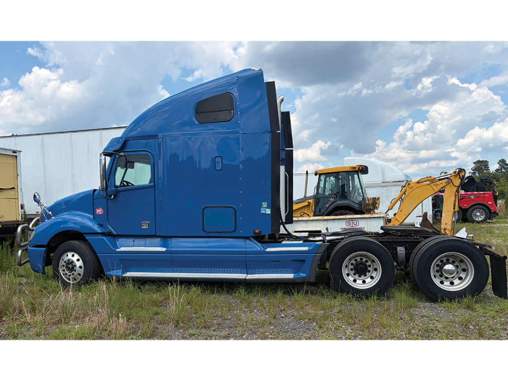 Freightliner truck tractor in blue, side view showing sleeper cab and dual rear axles with attached trailer hitch equipment.