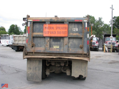 Dump truck rear showing Material Spreader sign and plate number 128, with mud, dual rear wheels and hitch. Visible text: MATERIAL SPREADER KEEP BACK 300 FEET.