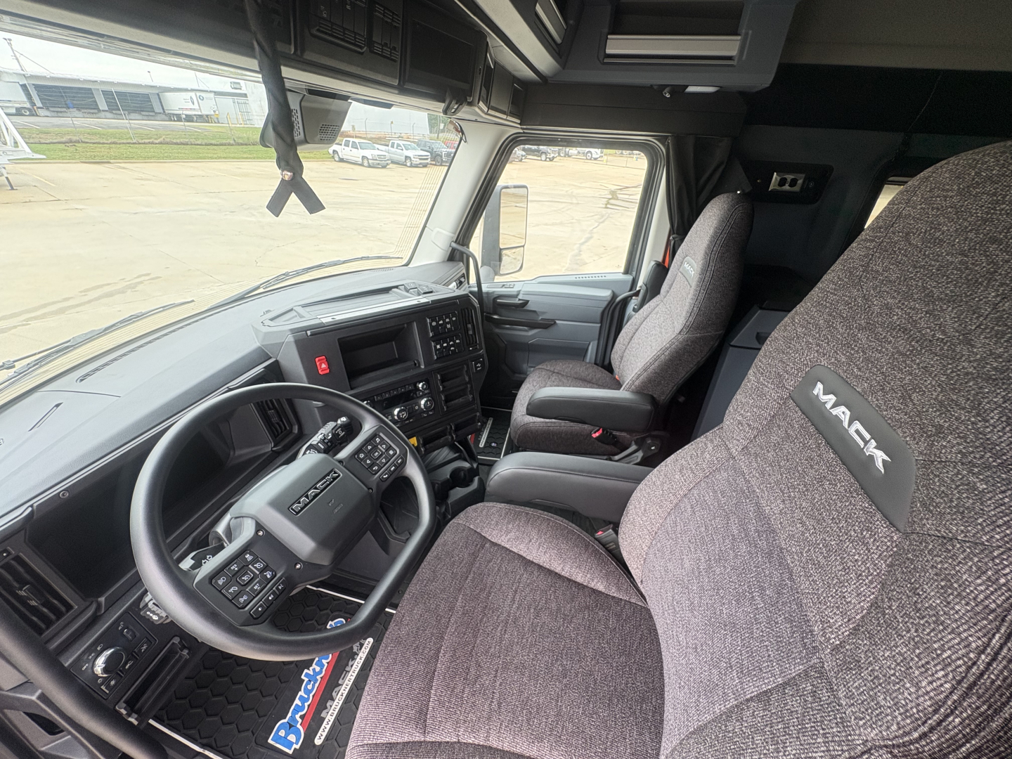 Truck cab interior showing steering wheel, dashboard, and two gray fabric seats with Mack branding on headrest in a new Mack AN64T64 conventional daycab.