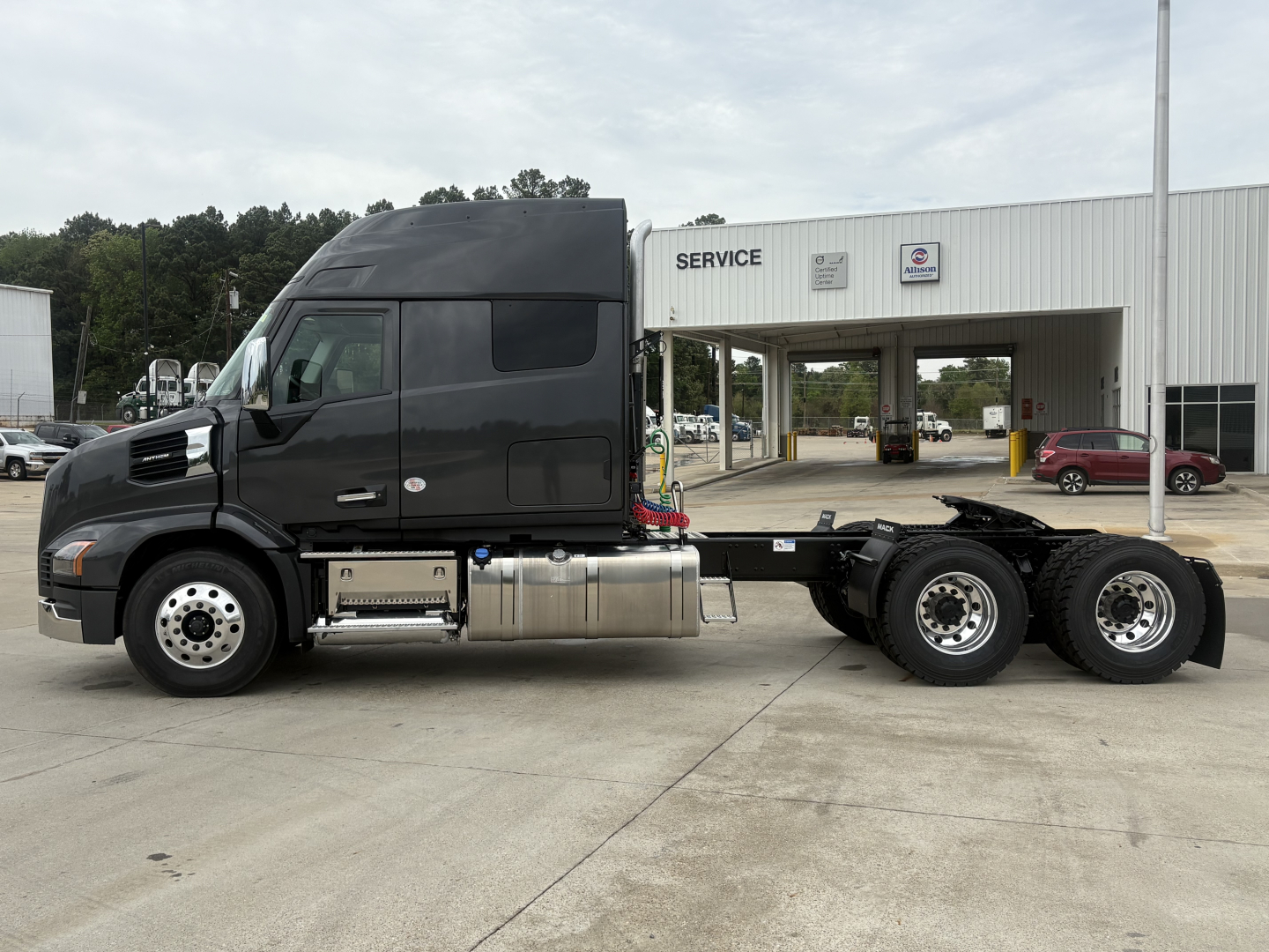 Truck chassis side view showing 6x4 drivetrain on a new 2027 Mack AN64T64 conventional daycab, with cab high roof, fuel tanks, and service building in background.