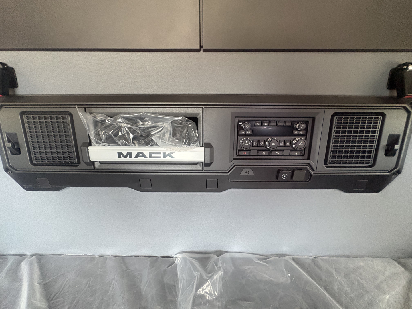 Dashboard area of a Mack AN64T64 truck showing center console with radio, vents, and a Mack badge in a plastic bag. Visible text includes 'MACK' on the badge and control panel markings.