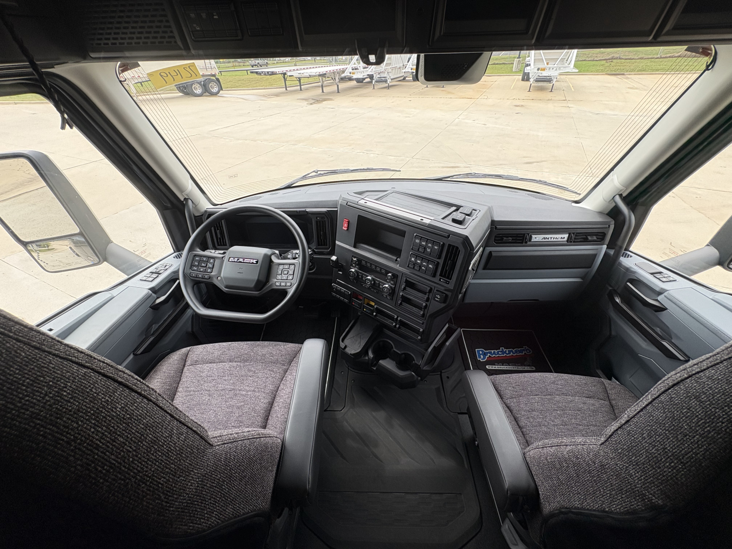 Cab interior of a Mack AN64T64 daycab showing steering wheel, dashboard with radio, center console, and two cloth seats.