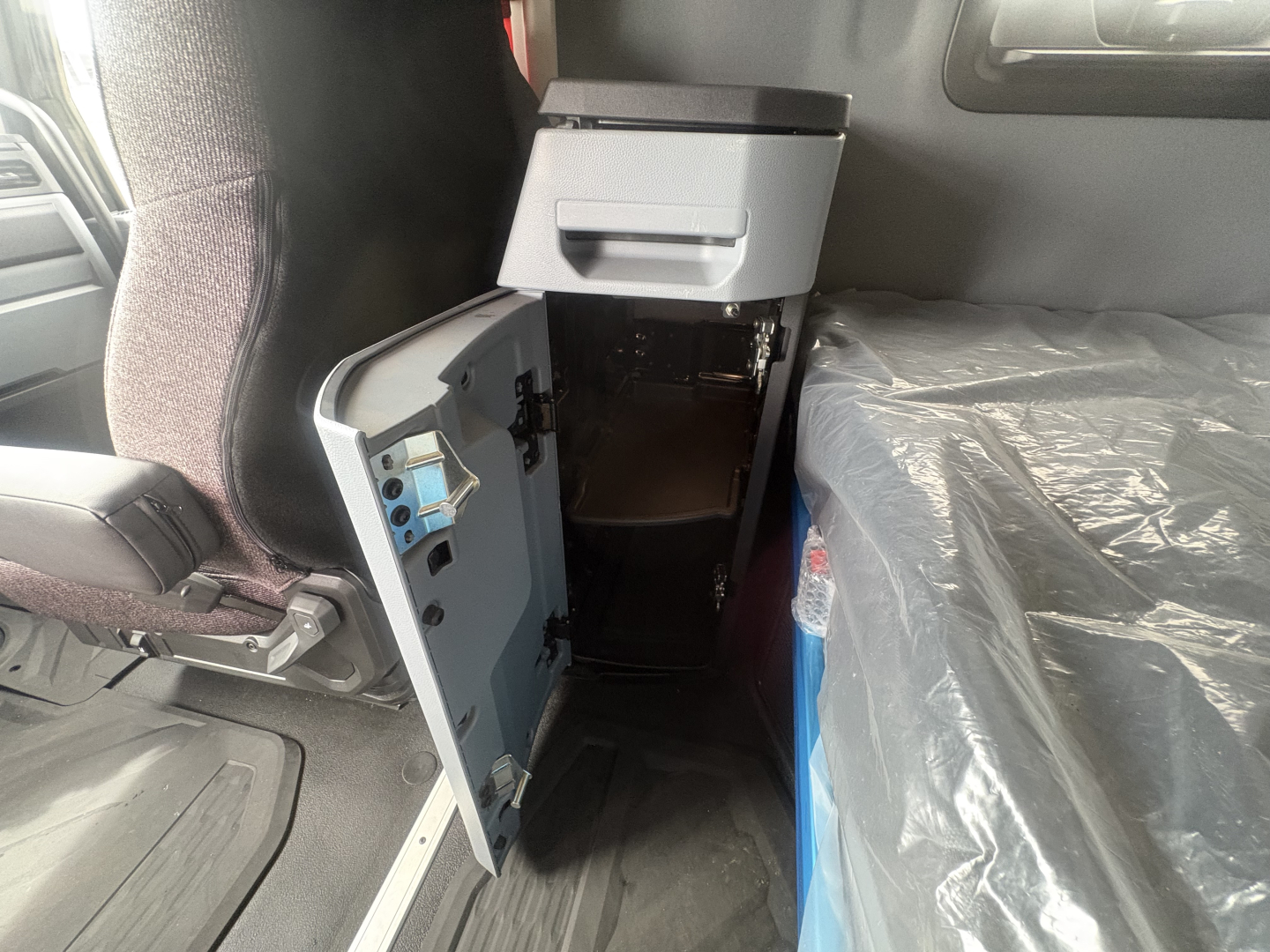 Cab interior of a 2027 Mack AN64T64 conventional daycab showing an open storage compartment with blue painted interior and a plastic-covered seat or bench to the right.