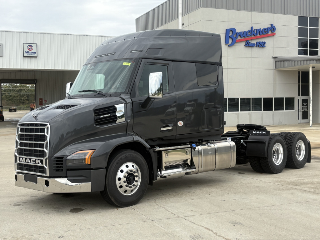 Mack AN64T64 conventional daycab truck parked outside dealership showing chrome fuel tanks and dual rear axles.