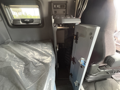 Cab interior of a new 2027 Mack AN64T64 truck showing a small open cabinet/locker with a door open, control panel above, and plastic-covered seat.