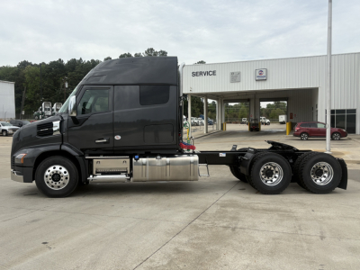 Truck chassis side view showing 6x4 drivetrain on a new 2027 Mack AN64T64 conventional daycab, with cab high roof, fuel tanks, and service building in background.
