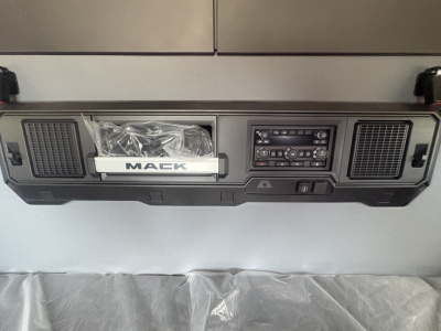 Dashboard area of a Mack AN64T64 truck showing center console with radio, vents, and a Mack badge in a plastic bag. Visible text includes 'MACK' on the badge and control panel markings.