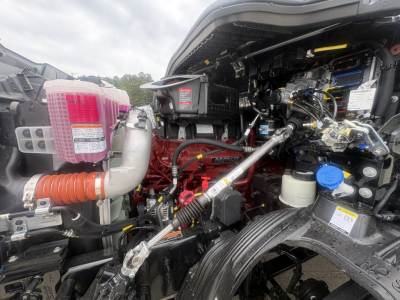 Truck engine bay of a new Mack AN64T64 with visible hoses, reservoirs, and wiring; pink coolant reservoir cap, blue cap, and various labels visible.