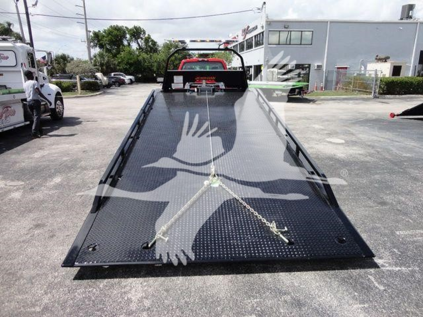 Flatbed tow truck ramp with diamond plate surface and metal wheel straps, front winch bar visible in background.