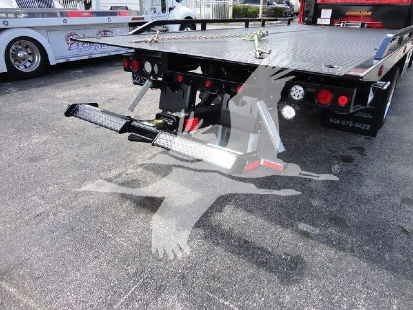 Flatbed tow truck rear with hydraulic winch and ramps, pipe controls, and reflective decals on a trailer; clear view of platform and hitch area.