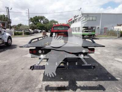 Flatbed rollback tow truck chassis with rear loading ramps and hydraulic system, viewed from behind in a dealership lot.