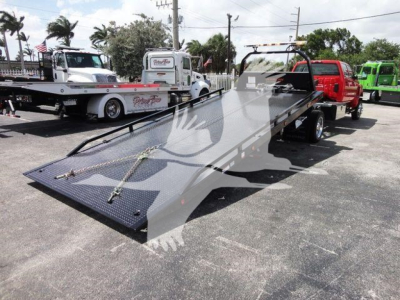 Tow truck rollback bed with hydraulic flatbed in a parking lot, black surface, metal chains and ramp extensions visible.