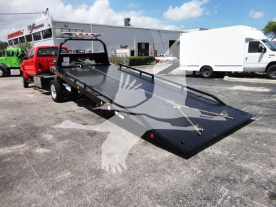 Flatbed rollback tow truck on a lot with other trucks and a white van in the background; tilted downbed showing metallic deck and tie-down anchors.
