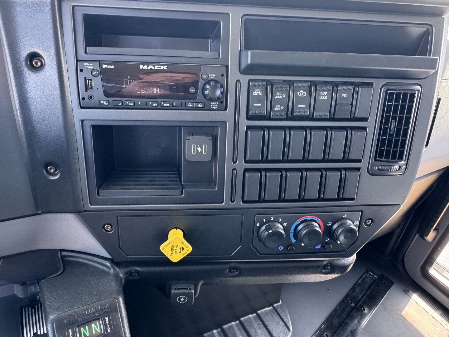 Mack MD7 dash and center console showing radio, numerous switch panels, climate controls, and a yellow hood release knob.