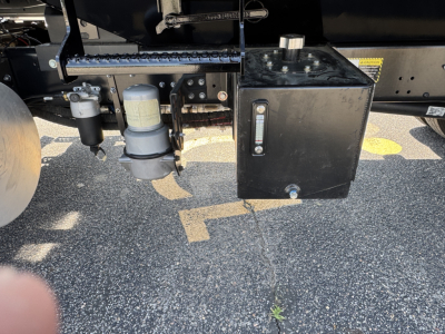 Underbelly of a 2025 Mack MD7 truck showing a black rectangular toolbox with a vertical gauge, a gray canister, and a hydraulic/air line assembly connected to the frame.