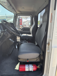 Truck cab interior of 2025 Mack MD7 showing driver seat and passenger seat, steering wheel, dashboard, and a red fire extinguisher under the seat.