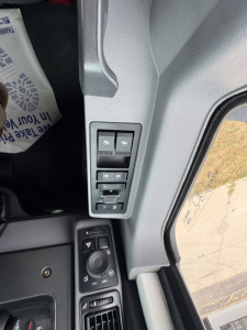 Truck cab interior showing window/mirror controls panel with multiple switches and a joystick-style knob, near a side window.