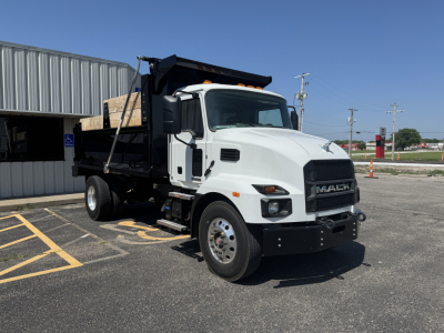 2025 Mack MD7 dump truck in a parking lot with white cab, black grille, and dump body. Visible text includes MACK logo on grille and various stickers on cab and dump.