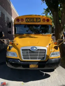 School bus front view showing bright yellow body, large grille, and windshield with 'SCHOOL BUS' sign; mirrors extended on both sides.