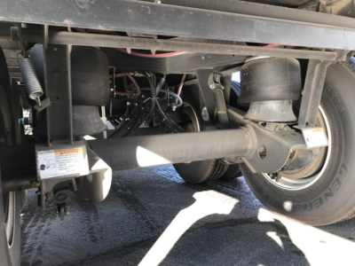 Trailer undercarriage showing axle, suspension, and dual wheels with tires; various hoses and wiring visible; label plate and mechanical components near frame