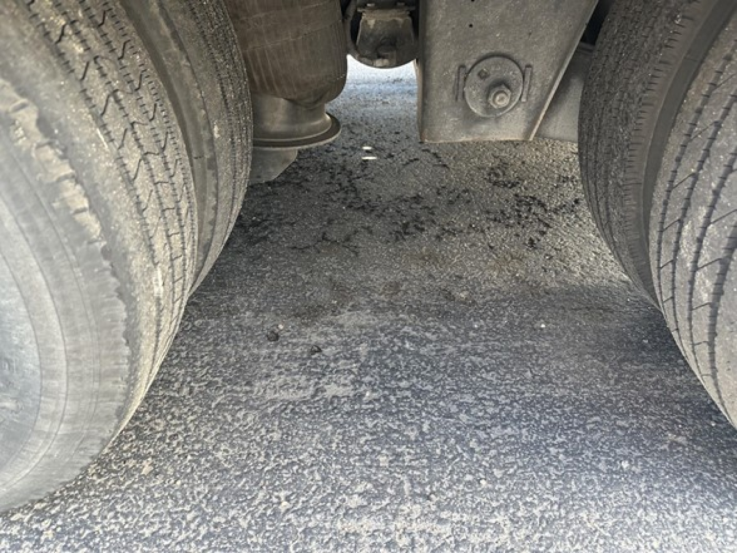 Trailer undercarriage showing dual rear axles with tires and a central wear plate; view between axles with ground surface visible.