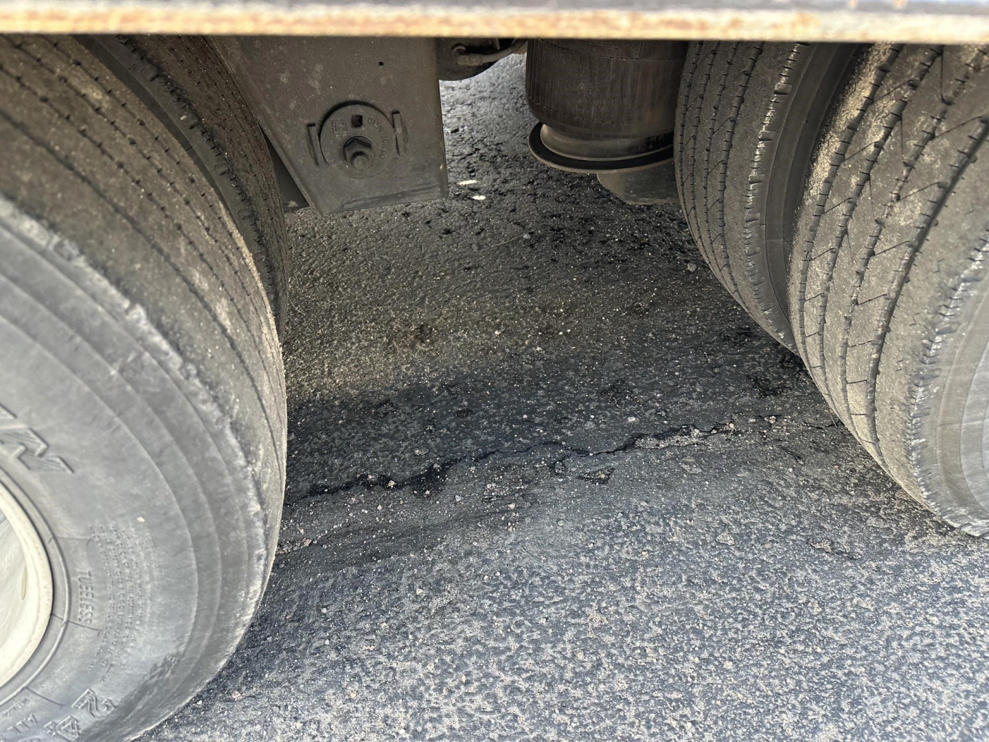 Trailer underside view showing dual rear wheels and suspension, with rough asphalt surface and debris between tires.