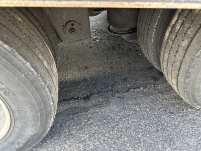 Trailer underside view showing dual rear wheels and suspension, with rough asphalt surface and debris between tires.