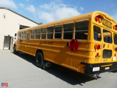School bus side and rear view showing yellow body, blue sky, school district label, and rear taillights.